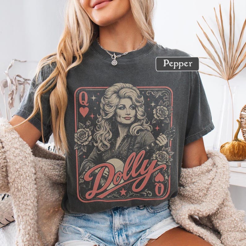 May include: A charcoal gray short-sleeve t-shirt featuring a graphic print of a woman with voluminous hair playing a guitar. The design includes roses, stars, heart symbols, the letter Q, and the word Dolly in a script font. The text Pepper is visible.