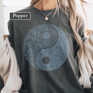 Puede incluir: Sudadera gris oscuro con un diseño de yin yang azul lleno de patrones florales y estrellas. La palabra "Pepper" está en un rectángulo blanco. La sudadera tiene cuello redondo.