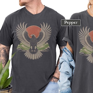 May include: Dark gray t-shirts featuring a graphic of an eagle with outstretched wings, a red sun, and green mountains. The word "Pepper" is in a white box. The shirts have a vintage, worn look.