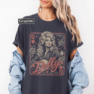 May include: A graphite-colored t-shirt featuring a graphic print of a woman with blonde hair playing a banjo, framed like a playing card with the letter Q, roses, and stars. The text Dolly is printed in a large, stylized red script font across the front.