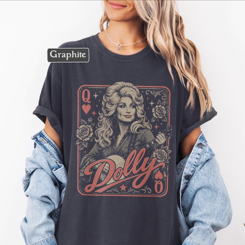 May include: A graphite-colored t-shirt featuring a graphic print of a woman with blonde hair playing a banjo, framed like a playing card with the letter Q, roses, and stars. The text Dolly is printed in a large, stylized red script font across the front.