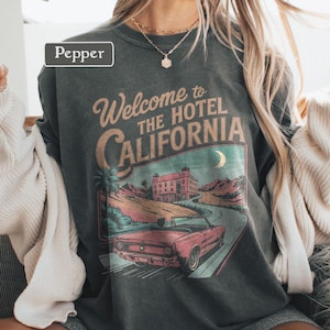 May include: Dark gray t-shirt with a graphic that reads "Welcome to The Hotel California." The graphic features a vintage car, a hotel, palm trees, and a crescent moon. The shirt has a vintage, worn look.