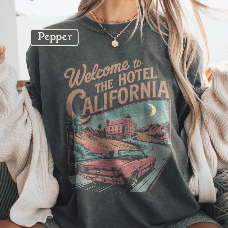 May include: Dark gray t-shirt with a graphic that reads "Welcome to The Hotel California." The graphic features a vintage car, a hotel, palm trees, and a crescent moon. The shirt has a vintage, worn look.
