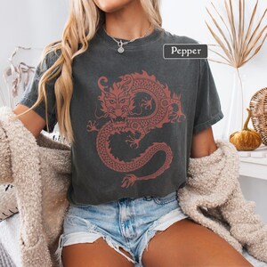 May include: Dark gray t-shirt featuring a red dragon design. The dragon is intricately detailed, with a long, winding body and a fierce expression. The word "Pepper" is in a white rectangle on the top right of the shirt.