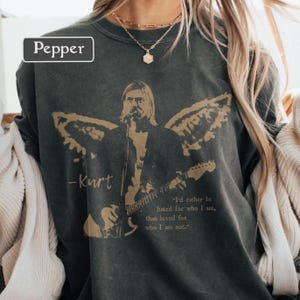 May include: Dark gray sweatshirt featuring a gold graphic of a musician playing a guitar with angel wings. The text "-Kurt" and a quote, "I'd rather be hated for who I am, than loved for who I am not," are also visible. The word "Pepper" is in a white box.