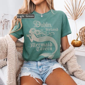 May include: Light green t-shirt featuring the text "Dublin, Ireland Mermaid Tavern" and a mermaid illustration. The shirt has a vintage aesthetic, with a faded light green hue. The text and graphic are in a light beige colour.