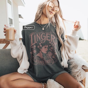 May include: Dark gray sweatshirt featuring the text "Linger" and "The Cranberries" in a pink, vintage-style design. The design includes a portrait of a woman surrounded by roses. The sweatshirt is worn with a white cardigan and gray shorts.