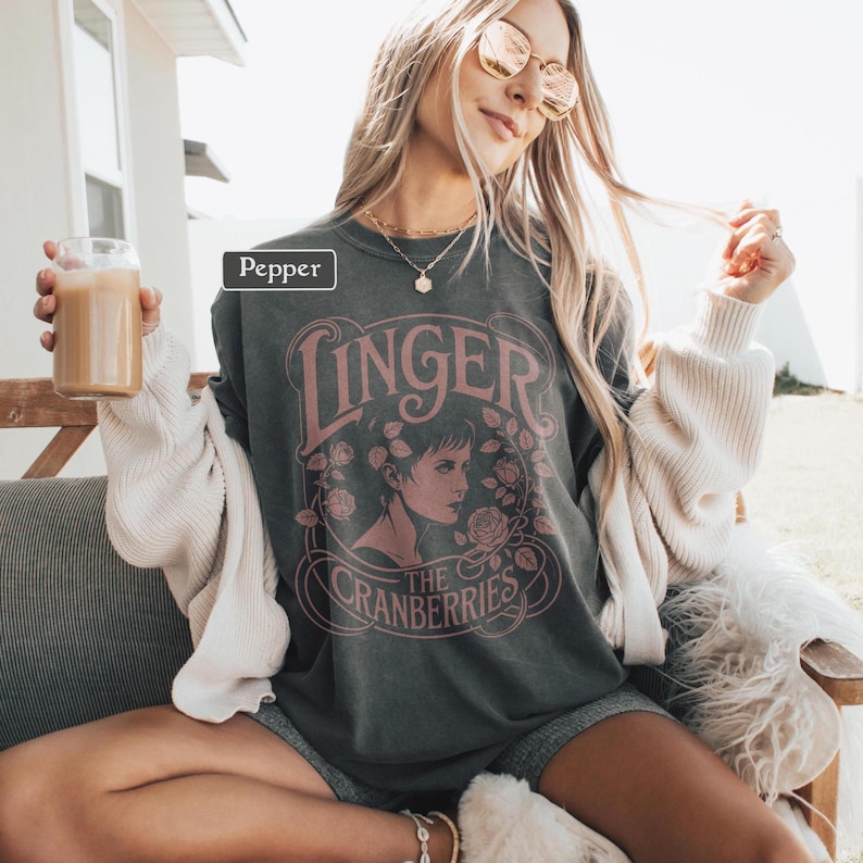 May include: Dark gray sweatshirt featuring the text "Linger" and "The Cranberries" in a pink, vintage-style design. The design includes a portrait of a woman surrounded by roses. The sweatshirt is worn with a white cardigan and gray shorts.