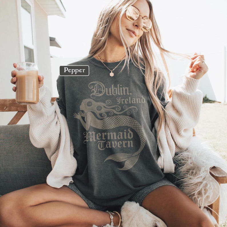 May include: A dark grey t-shirt featuring the text "Dublin, Ireland Mermaid Tavern" and a mermaid illustration. The shirt has a relaxed fit and a vintage appearance. The model is wearing sunglasses and holding a glass of a beverage.
