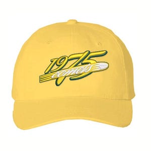 May include: Yellow baseball cap with the text "1975 comets" in a stylized design. The text is in yellow with a white and green outline. The cap is a solid yellow color.