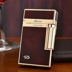 May include: A rectangular, brown lighter with gold accents. The lighter has the word "Derui" and the word "Lighter" printed on it. The lighter has a gold-colored, ribbed side and a gold-colored frame.