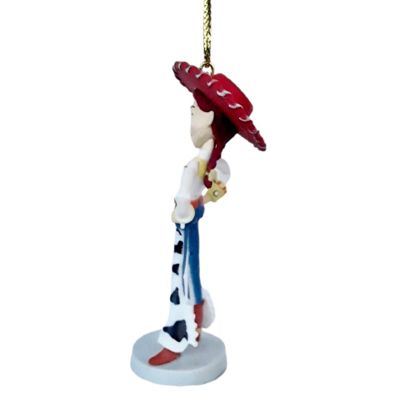 May include: A Jessie figurine from the Toy Story franchise. The figurine is wearing a red hat, white shirt, blue pants with cow print, and red boots. The figurine is attached to a gold string for hanging.