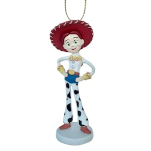 May include: A Jessie ornament from the Toy Story franchise. The ornament features Jessie in her signature cowgirl outfit, including a red hat, white shirt, blue shorts, and cow-print pants. The ornament is attached to a gold-colored string.