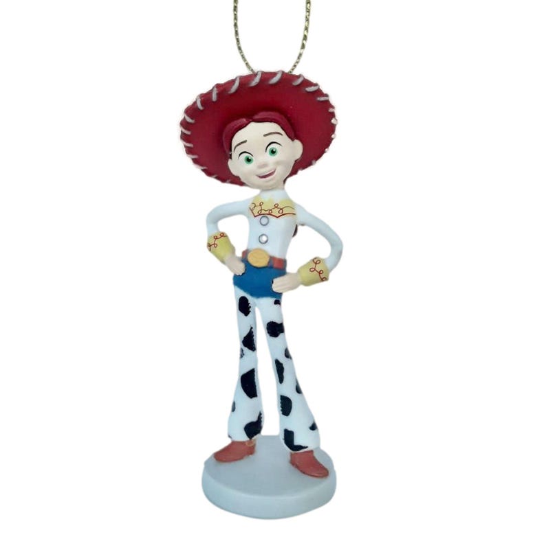 May include: A Jessie ornament from the Toy Story franchise. The ornament features Jessie in her signature cowgirl outfit, including a red hat, white shirt, blue shorts, and cow-print pants. The ornament is attached to a gold-colored string.