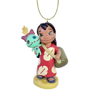 Pode incluir: Um enfeite decorativo com um personagem do filme de anima&ccedil;&atilde;o Lilo & Stitch. A figura &eacute; de uma menina em um vestido vermelho com desenhos florais brancos, segurando uma boneca azul-turquesa. O enfeite &eacute; suspenso por uma corrente dourada.