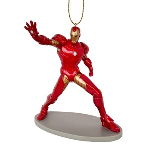 May include: A red and gold Iron Man figurine ornament. The superhero figure is in a dynamic pose, with one arm outstretched. The ornament has a gold string for hanging and is mounted on a gray oval base. The figure is approximately 5 inches tall.