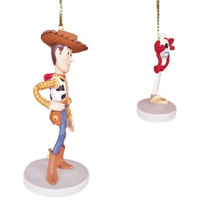 May include: Two Christmas ornaments featuring characters from the Toy Story franchise. The first ornament is a figurine of Woody, the cowboy doll, wearing his signature brown hat, red bandana, and blue jeans. The second ornament is a figurine of Buzz Lightyear, the space ranger, with his signature red and white spacesuit and helmet. Both ornaments are hanging from gold strings.