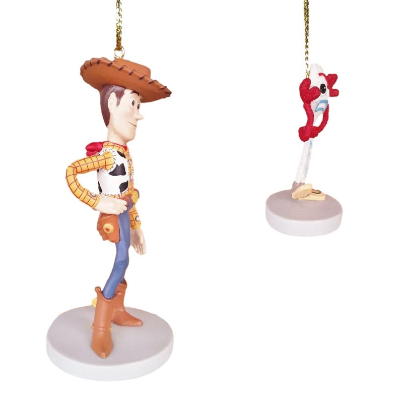 May include: Two Christmas ornaments featuring characters from the Toy Story franchise. The first ornament is a figurine of Woody, the cowboy doll, wearing his signature brown hat, red bandana, and blue jeans. The second ornament is a figurine of Buzz Lightyear, the space ranger, with his signature red and white spacesuit and helmet. Both ornaments are hanging from gold strings.