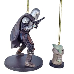 May include: A Mandalorian figure ornament with a silver helmet, black cape, and blaster, standing on a black base. Beside it, a small Grogu ornament in a tan cloth, also on a black base, hangs from a gold string.