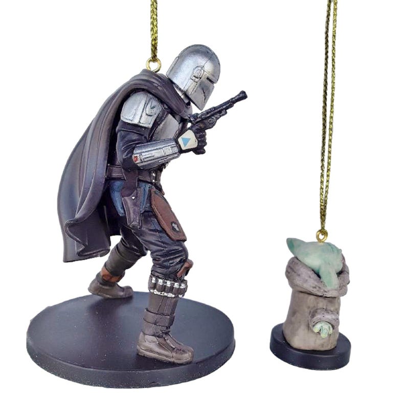May include: A Mandalorian figure ornament with a silver helmet, black cape, and blaster, standing on a black base. Beside it, a small Grogu ornament in a tan cloth, also on a black base, hangs from a gold string.