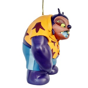 Op de afbeelding: Een beeldje van een stripfiguur met een geel shirt met kastanjebruine vormen, paarse armen en benen en een blauwe korte broek. Het personage heeft een bruin gezicht met een dreigende uitdrukking en een blauwe mond. Bovenop is een gouden touwtje bevestigd.