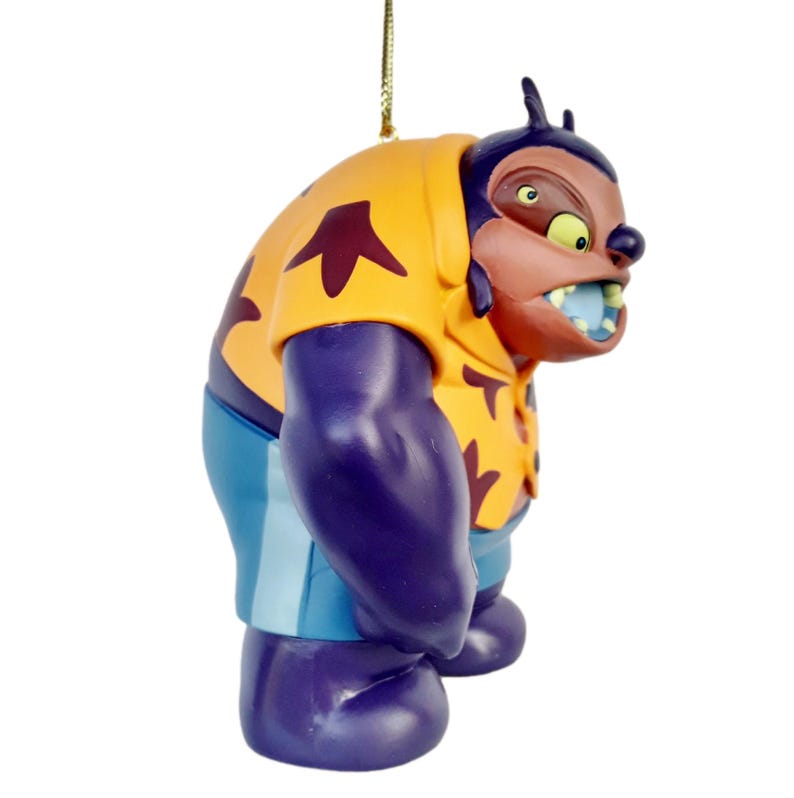 Op de afbeelding: Een beeldje van een stripfiguur met een geel shirt met kastanjebruine vormen, paarse armen en benen en een blauwe korte broek. Het personage heeft een bruin gezicht met een dreigende uitdrukking en een blauwe mond. Bovenop is een gouden touwtje bevestigd.