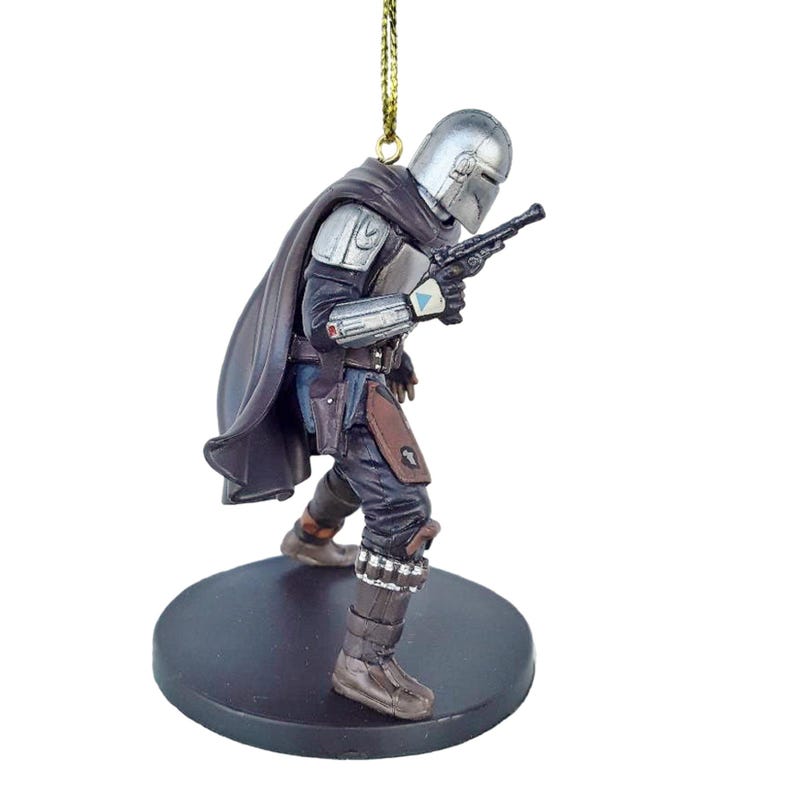 May include: A silver and brown Mandalorian ornament with a black cape and a blaster pistol. The ornament is standing on a black base.