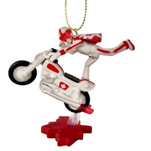 May include: A white and red Christmas ornament featuring a figure on a motorcycle performing a stunt. The figure is wearing a white jumpsuit with red accents and a red helmet. The motorcycle has a red Canadian flag on the side. The ornament is suspended from a gold string.