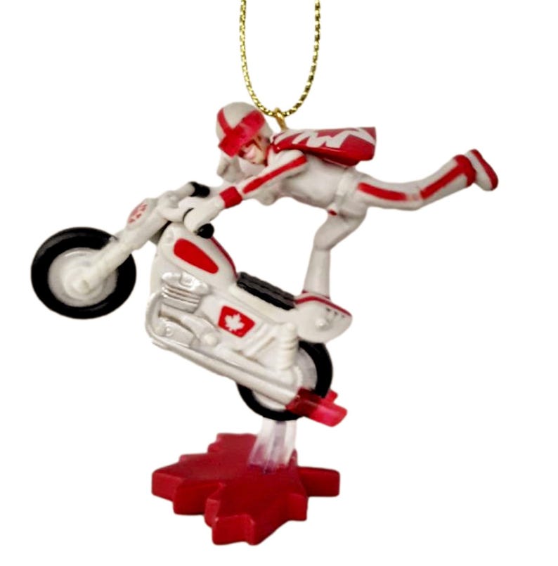 May include: A white and red Christmas ornament featuring a figure on a motorcycle performing a stunt. The figure is wearing a white jumpsuit with red accents and a red helmet. The motorcycle has a red Canadian flag on the side. The ornament is suspended from a gold string.