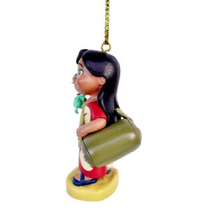 Pode incluir: Enfeite decorativo com um personagem de desenho animado em um vestido vermelho com uma bolsa verde. A figura tem cabelo preto e est&aacute; em p&eacute; sobre uma base bege. Um fio dourado &eacute; anexado para pendurar.