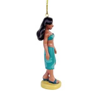 Peut inclure: Figurine d'une femme en haut de bikini et jupe-sarong turquoise, avec des cheveux noirs. La figurine porte des sandales bleues et se tient sur une base jaune. Une ficelle dor&eacute;e est attach&eacute;e au sommet.
