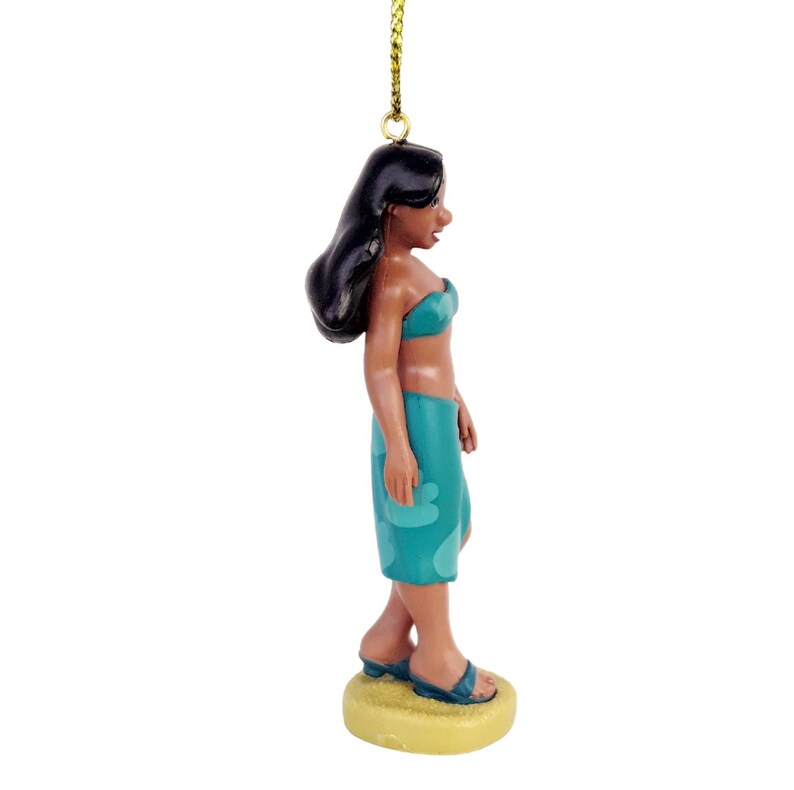 Peut inclure: Figurine d'une femme en haut de bikini et jupe-sarong turquoise, avec des cheveux noirs. La figurine porte des sandales bleues et se tient sur une base jaune. Une ficelle dor&eacute;e est attach&eacute;e au sommet.