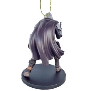 May include: A black and silver Mandalorian ornament with a dark purple cape. The ornament is standing on a black base and has a gold string for hanging.
