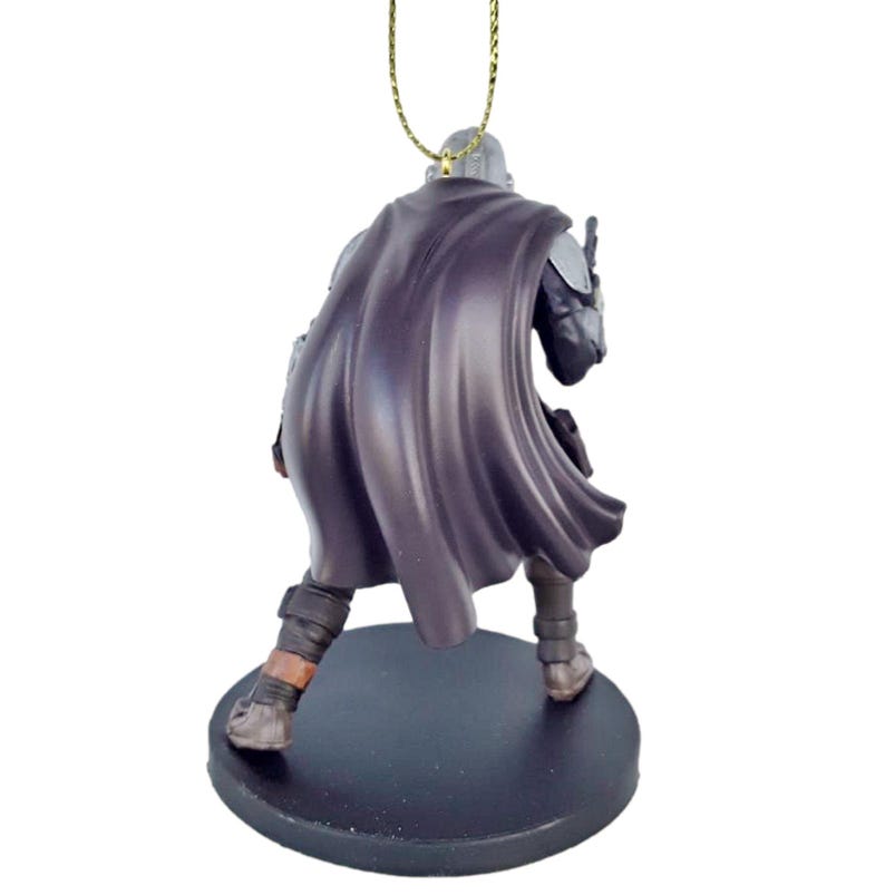 May include: A black and silver Mandalorian ornament with a dark purple cape. The ornament is standing on a black base and has a gold string for hanging.