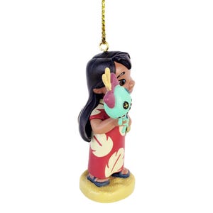 Pode incluir: Estatueta de uma menina em um vestido vermelho com estampas de folhas brancas, segurando uma boneca verde-&aacute;gua com cabe&ccedil;a amarela e roxa. A estatueta tem cabelo preto e est&aacute; em uma base bege. Uma corda dourada est&aacute; presa no topo.