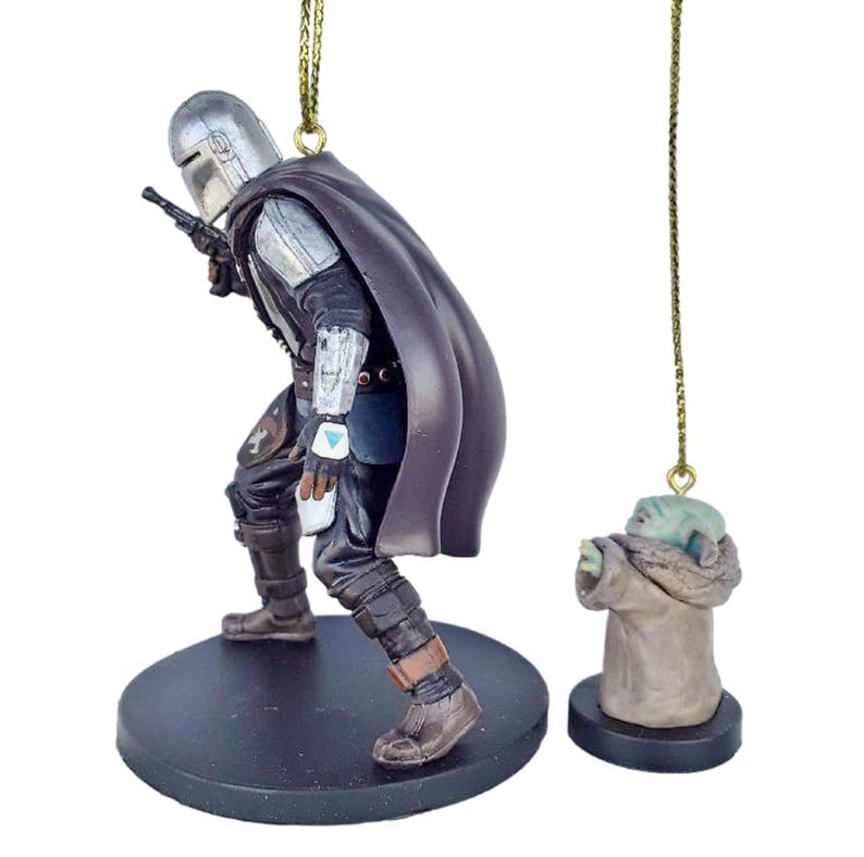May include: Two Star Wars-themed Christmas ornaments. One shows the Mandalorian in a black and silver suit with a blaster, and the other is a small figure of Grogu in a brown robe. Both ornaments have gold hanging strings.
