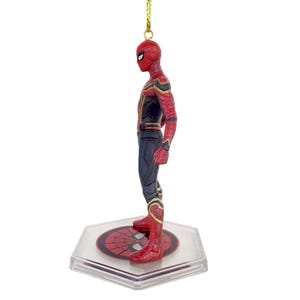May include: A Spider-Man figurine in a red and black suit with gold accents. The figure is standing on a clear, hexagonal base with a Spider-Man logo. A gold string is attached for hanging.