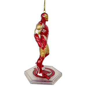 以下が含まれることがあります： 赤と金のアイアンマンのフィギュアオーナメント。スーパーヒーローのフィギュアは立っているポーズで、金色の頭とアクセントがあります。オーナメントは金の紐から吊り下げられ、赤いアベンジャーズのロゴが付いた透明なベースに取り付けられています。