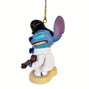 Puede incluir: Figura azul de Stitch vestido como Elvis Presley, con un mono blanco y una guitarra marr&oacute;n. La figura tiene pelo negro, una cuerda dorada y est&aacute; sobre una base beige. Sonr&iacute;e con los ojos cerrados.