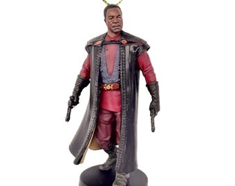 May include: A black and red Star Wars ornament featuring a character with a black cape and a red shirt. The character is holding a weapon in each hand.