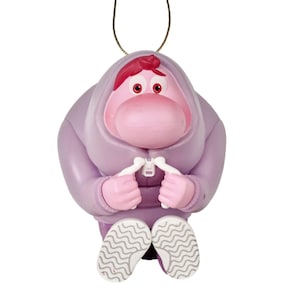May include: A purple ornament featuring a cartoon character with a pink face, red hair, and red eyes. The character wears a purple hooded sweatshirt and has white hands. The ornament has white and gray shoes and a gold string for hanging.