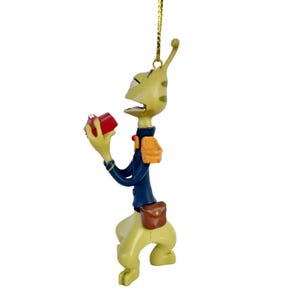 May include: A whimsical alien-like figurine ornament. The figure is light green with dark green spots, wearing a navy blue jacket, orange backpack, and brown satchel. It holds a small red box. The ornament hangs from a gold-colored string.