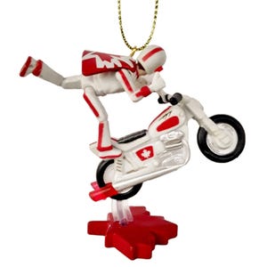 May include: A white and red ornament of a stunt motorcyclist performing a wheelie on a motorcycle. The motorcycle has a Canadian flag on the side. The ornament is attached to a gold string.
