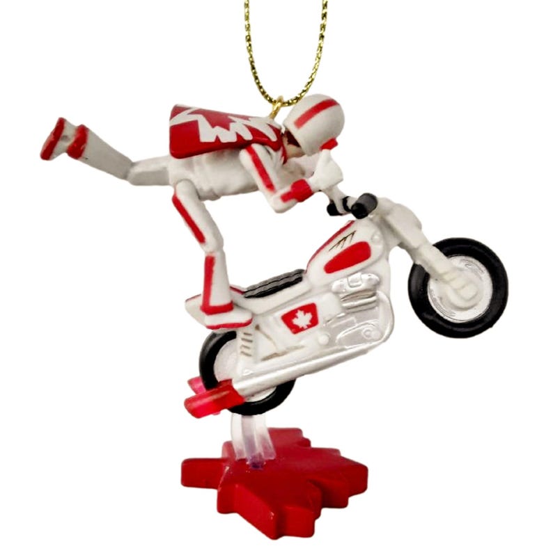 May include: A white and red ornament of a stunt motorcyclist performing a wheelie on a motorcycle. The motorcycle has a Canadian flag on the side. The ornament is attached to a gold string.