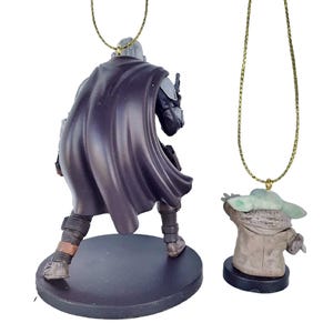May include: Two character ornaments from the Star Wars franchise. One is a figure of the Mandalorian in a dark cape, the other is a figure of Grogu, also known as Baby Yoda. Both figures are attached to gold-coloured chains.