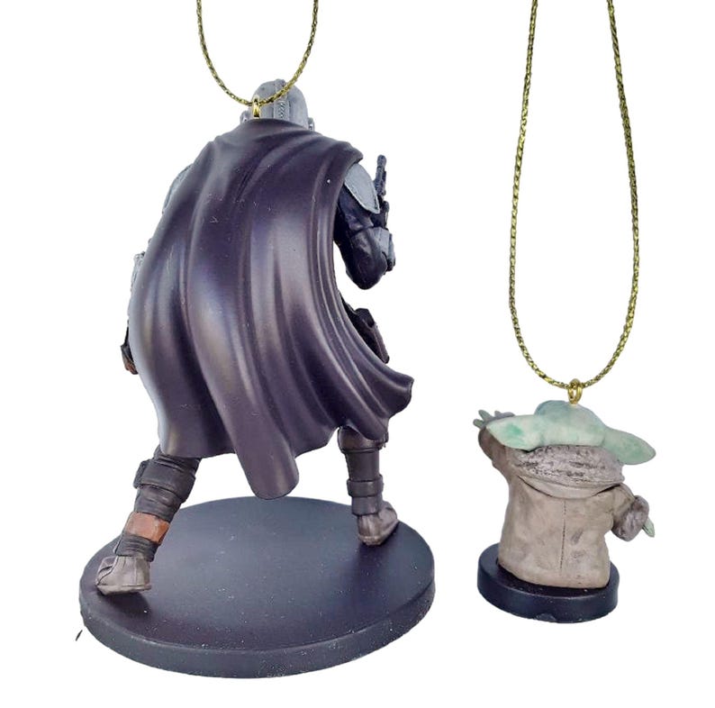 May include: Two character ornaments from the Star Wars franchise. One is a figure of the Mandalorian in a dark cape, the other is a figure of Grogu, also known as Baby Yoda. Both figures are attached to gold-coloured chains.