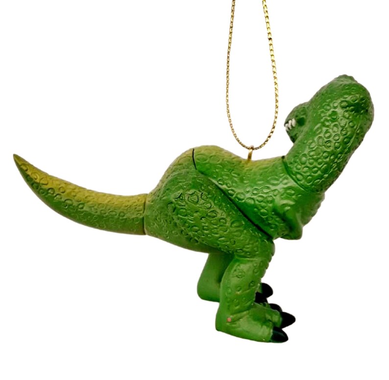 Puede incluir: Adorno de dinosaurio de pl&aacute;stico verde con una cuerda dorada para colgar. El dinosaurio est&aacute; en posici&oacute;n de cuclillas con la cabeza vuelta hacia atr&aacute;s.