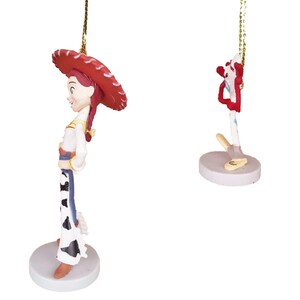 May include: Two Christmas ornaments featuring characters from the Toy Story franchise. The first ornament is a red-haired female character wearing a red and white cowboy hat and a white and black cowgirl outfit. The second ornament is a red and white character with a long, thin body and a blue and white striped shirt.