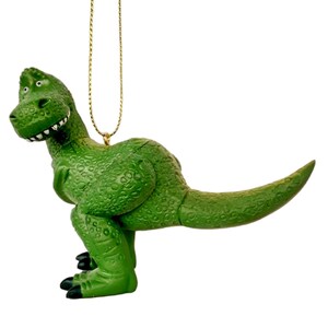 Puede incluir: Adorno de dinosaurio de pl&aacute;stico verde con una cuerda dorada para colgar. El dinosaurio est&aacute; de pie sobre sus cuatro patas con la boca abierta y la lengua fuera.
