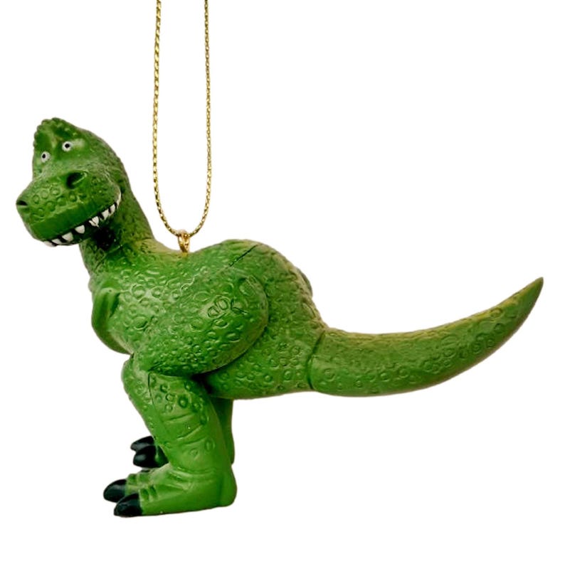 Puede incluir: Adorno de dinosaurio de pl&aacute;stico verde con una cuerda dorada para colgar. El dinosaurio est&aacute; de pie sobre sus cuatro patas con la boca abierta y la lengua fuera.
